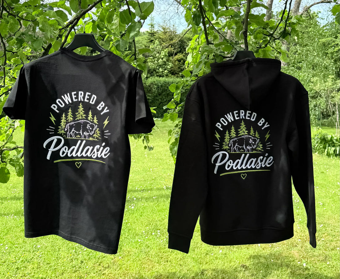 Czarna bluza z kapturem i czarna koszulka z haftem komputerowym "Powered by Podlasie" z motywem żubra i świerków, sfotografowane na tle zieleni